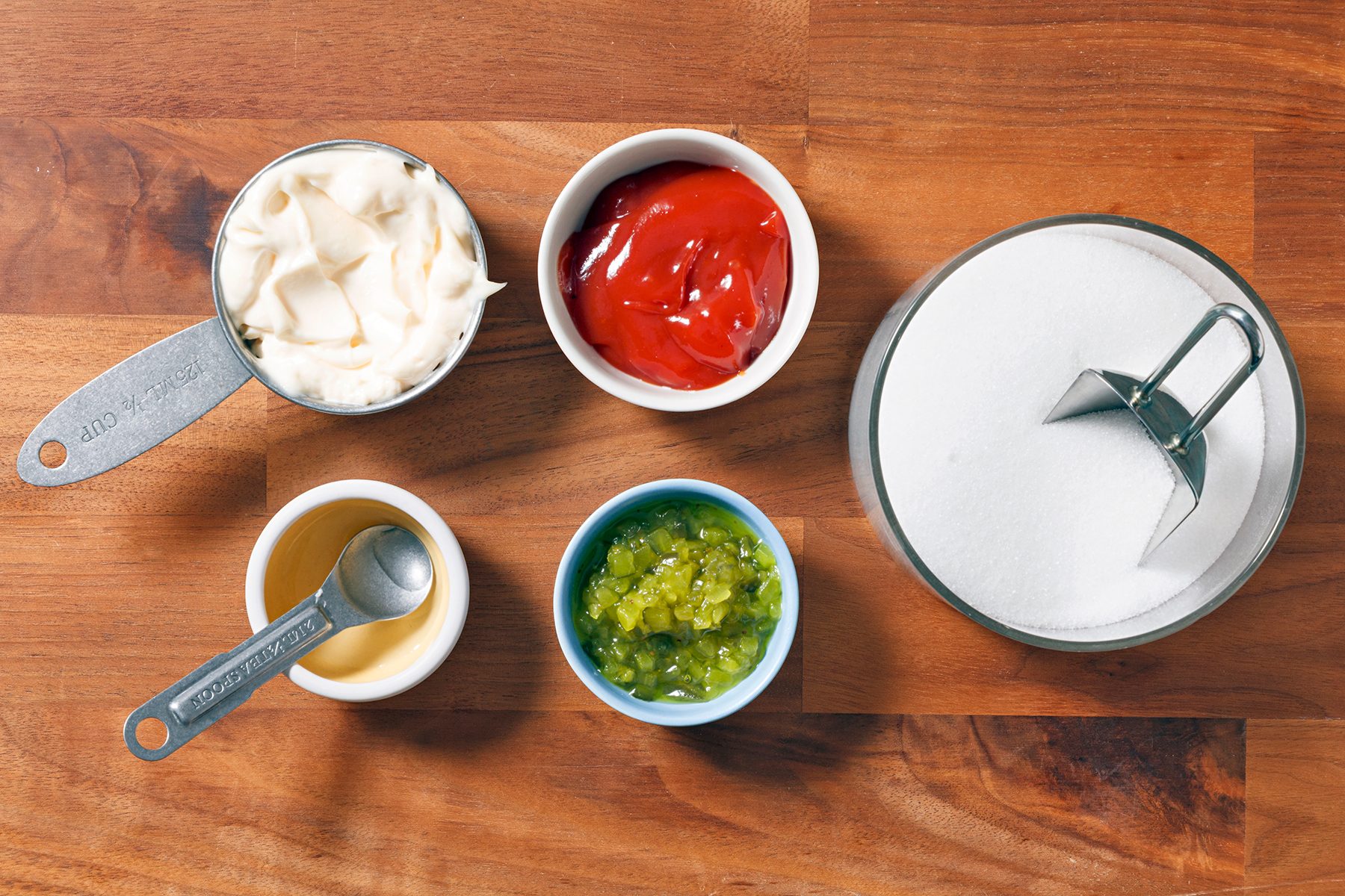 Four small bowls sit on a wooden surface containing mayonnaise, ketchup, sweet relish, and sugar. A measuring spoon and a cup are nearby.