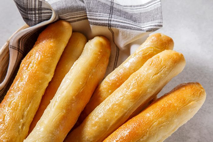 A basket of golden-brown breadsticks, lightly glazed, rests on a gray surface. The breadsticks are wrapped in a black and white checkered cloth.