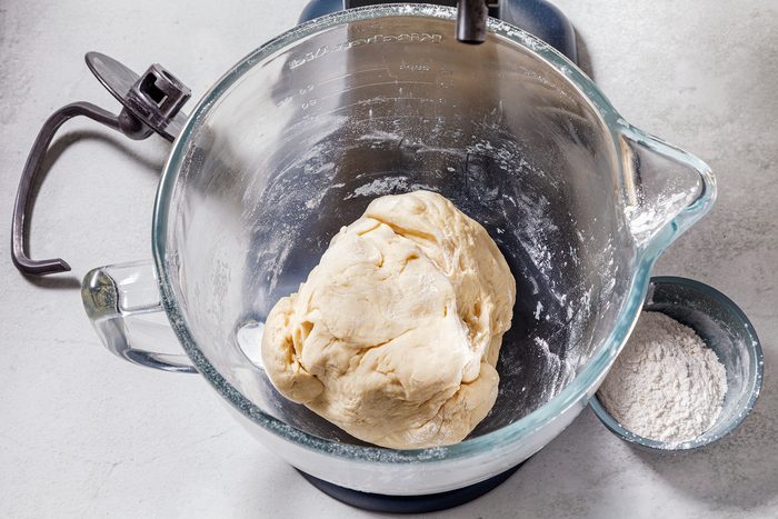 A glass mixing bowl with a ball of dough inside sits on a white countertop. Nearby, a small bowl holds some flour. A metal handle is attached to the bowl, suggesting it is part of a stand mixer.