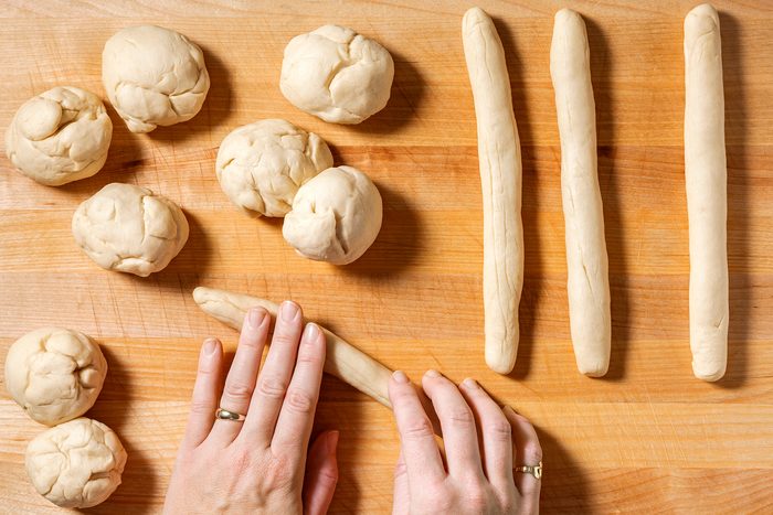 Hands shaping dough into long rolls on a wooden surface, with several dough balls nearby.