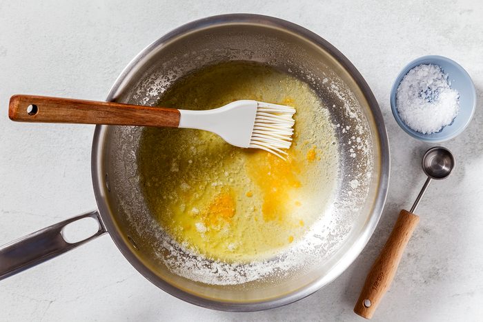 A saucepan with melted butter and a basting brush on a light surface. A small blue bowl with coarse salt and a wooden-handled measuring spoon are beside the pan.
