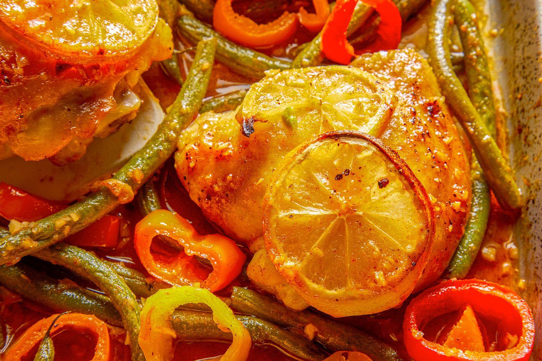 Close-up of a baked chicken thigh topped with lemon slices, surrounded by green beans, and sliced red and yellow bell peppers, all seasoned with spices.