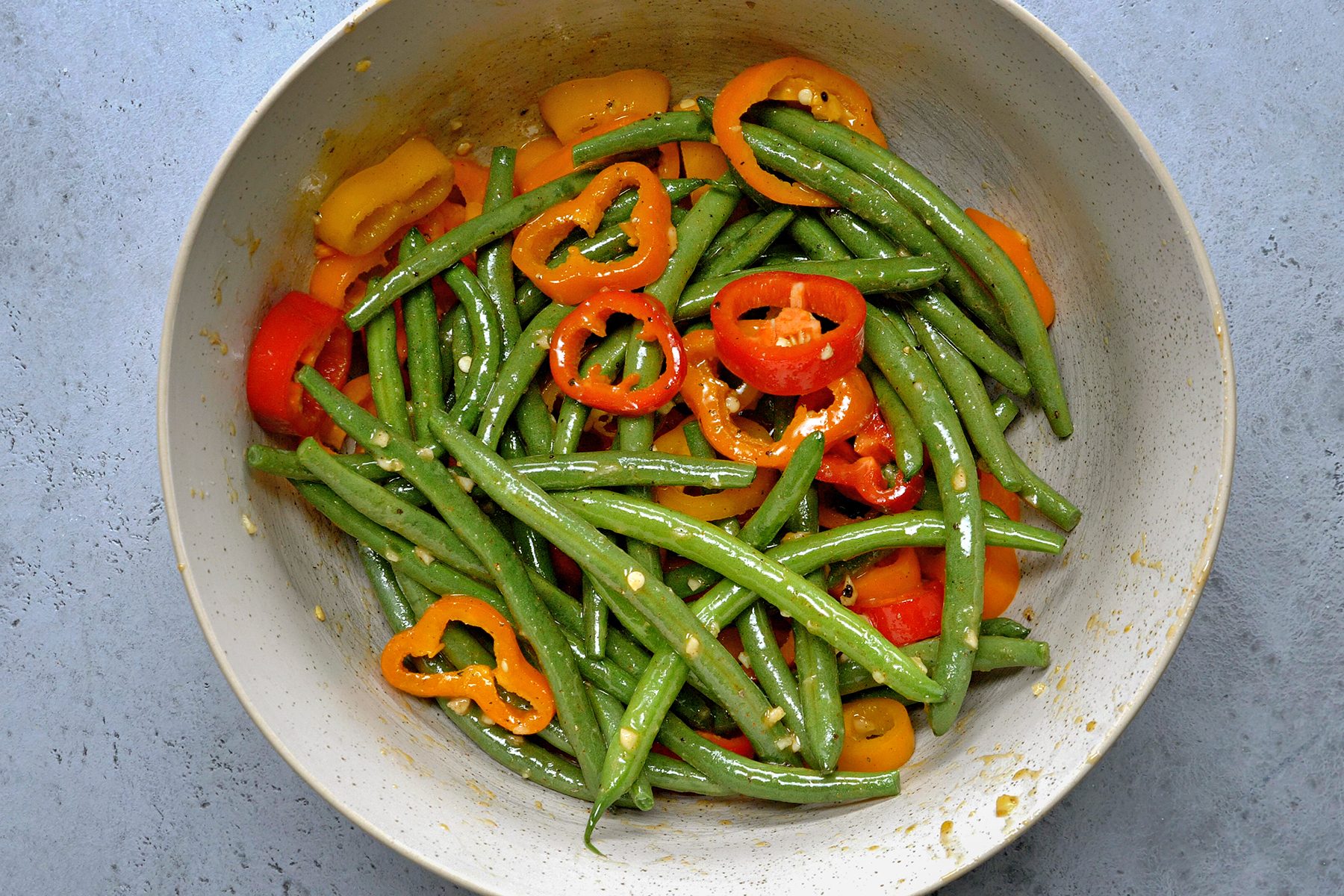 A bowl filled with green beans and sliced red, yellow, and orange bell peppers, all lightly seasoned. The dish is set against a gray background.