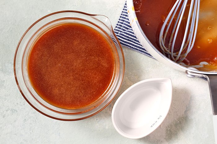A glass bowl filled with brown sauce sits on a light surface next to a small, empty white dish. A saucepan with sauce and a whisk is partially visible, resting on a blue and white striped cloth.