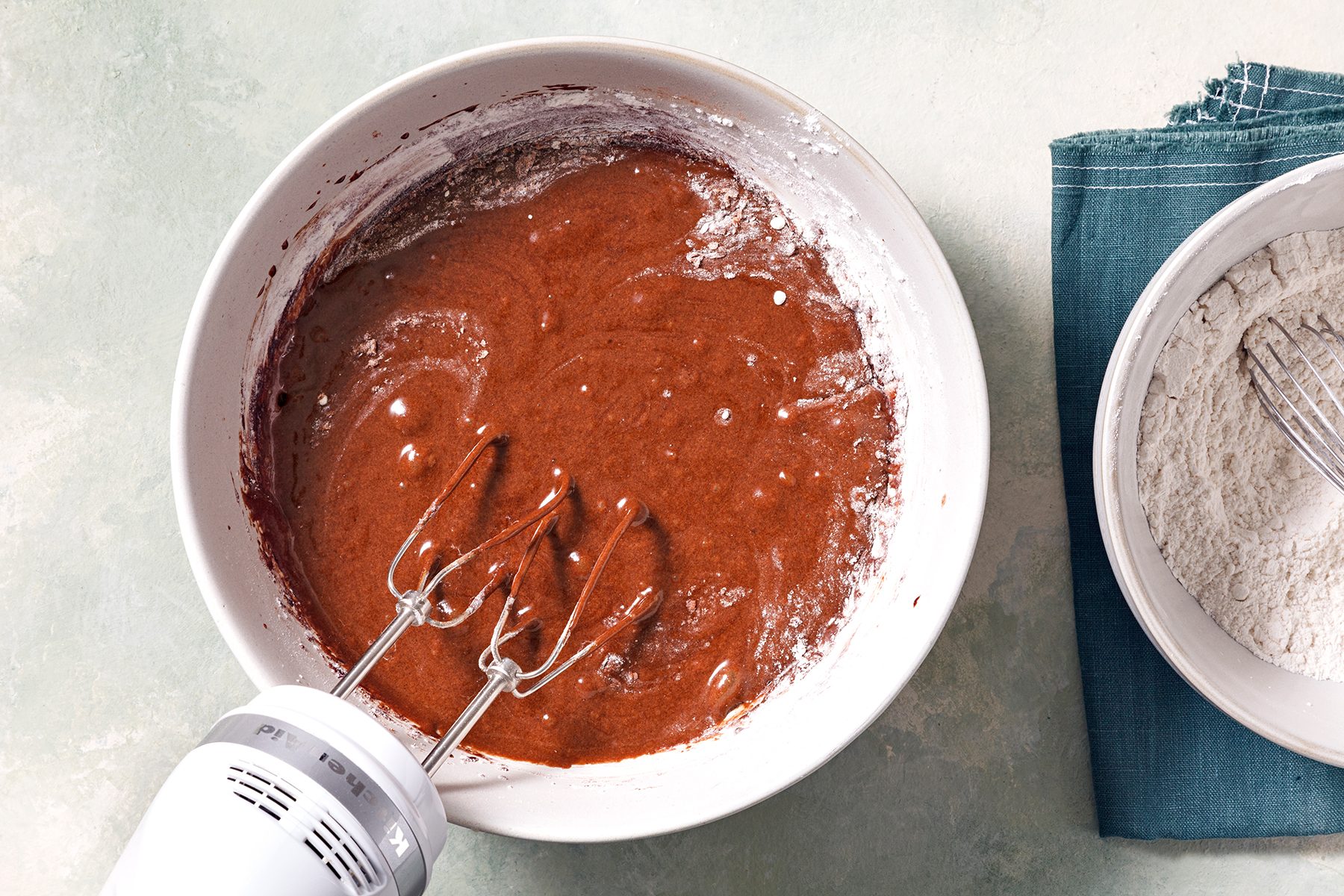 A bowl of chocolate cake batter being mixed with an electric hand mixer. Next to it is a bowl of flour with a whisk on a teal cloth. The surface is light-colored, creating a bright kitchen setting.