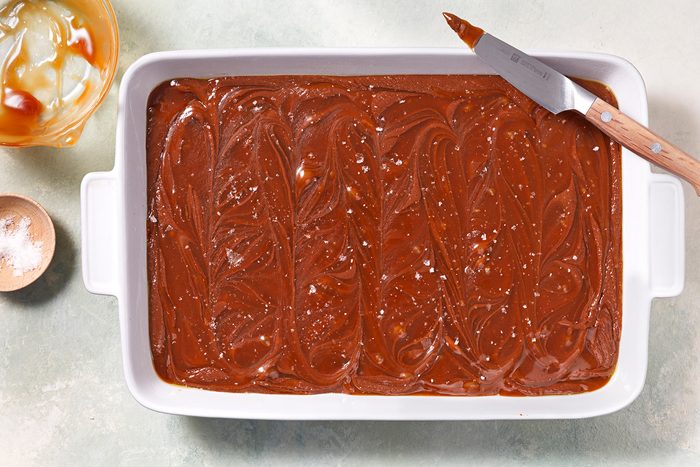A white rectangular dish filled with smooth, swirled chocolate-caramel fudge. A knife with chocolate residue rests on the dish's edge. Nearby, an empty spoon and a small bowl with caramel sauce are visible on a light surface.