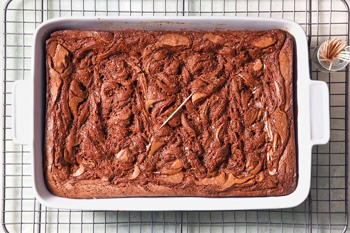 A rectangular baking dish filled with swirled chocolate brownies cooling on a wire rack. A toothpick is inserted in the center for doneness testing, and a small container of additional toothpicks is nearby.