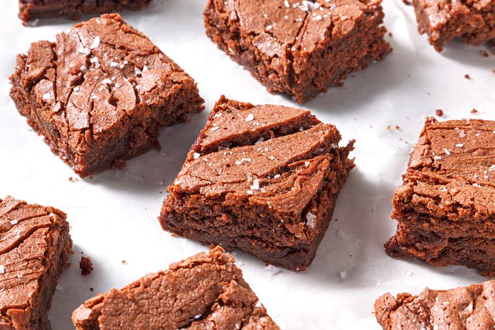 Squares of chocolate brownies sprinkled with sea salt are arranged on a white parchment paper. The brownies have a rich, fudgy texture with slightly cracked tops.