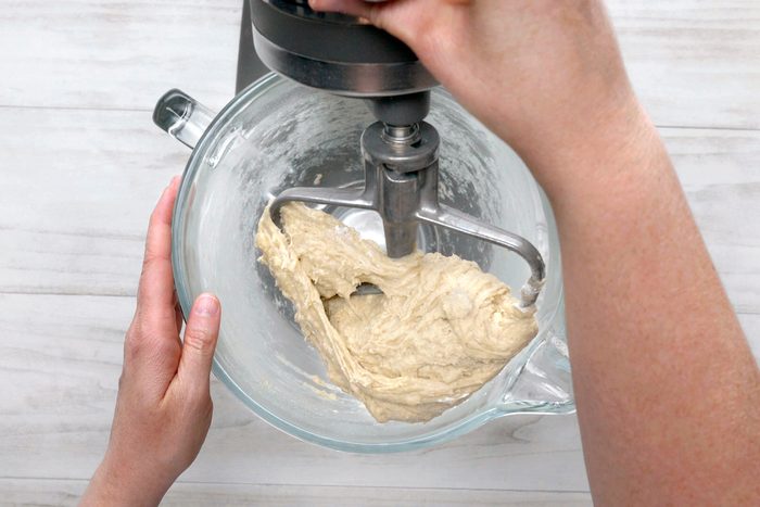overhead shot of a stand mixer with dough mixing inside; the mixer is in a glass bowl and the dough is a light brown color; a person is holding the bowl with their left hand and the right hand is turning the mixer