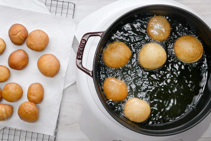 overhead shot of a black cast iron pan with a red handle sits on a white burner, filled with hot oil in which six round, golden-brown donuts are frying; on the left side of the image are twelve donuts of the same type, already fried and laid out on a white paper towel, with a wire cooling rack in the background