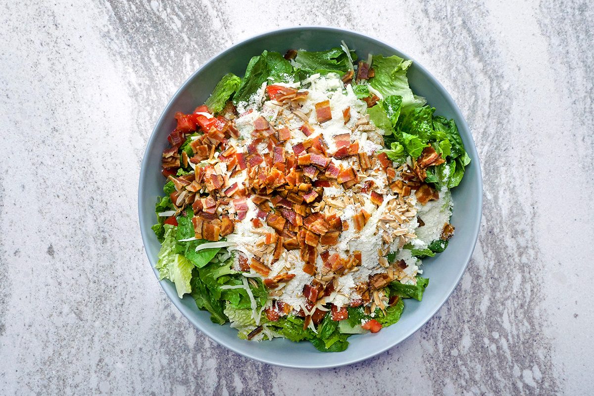 A bowl of chopped salad with fresh greens, diced tomatoes, shredded cheese, crispy bacon bits, and a creamy dressing on a marbled surface.