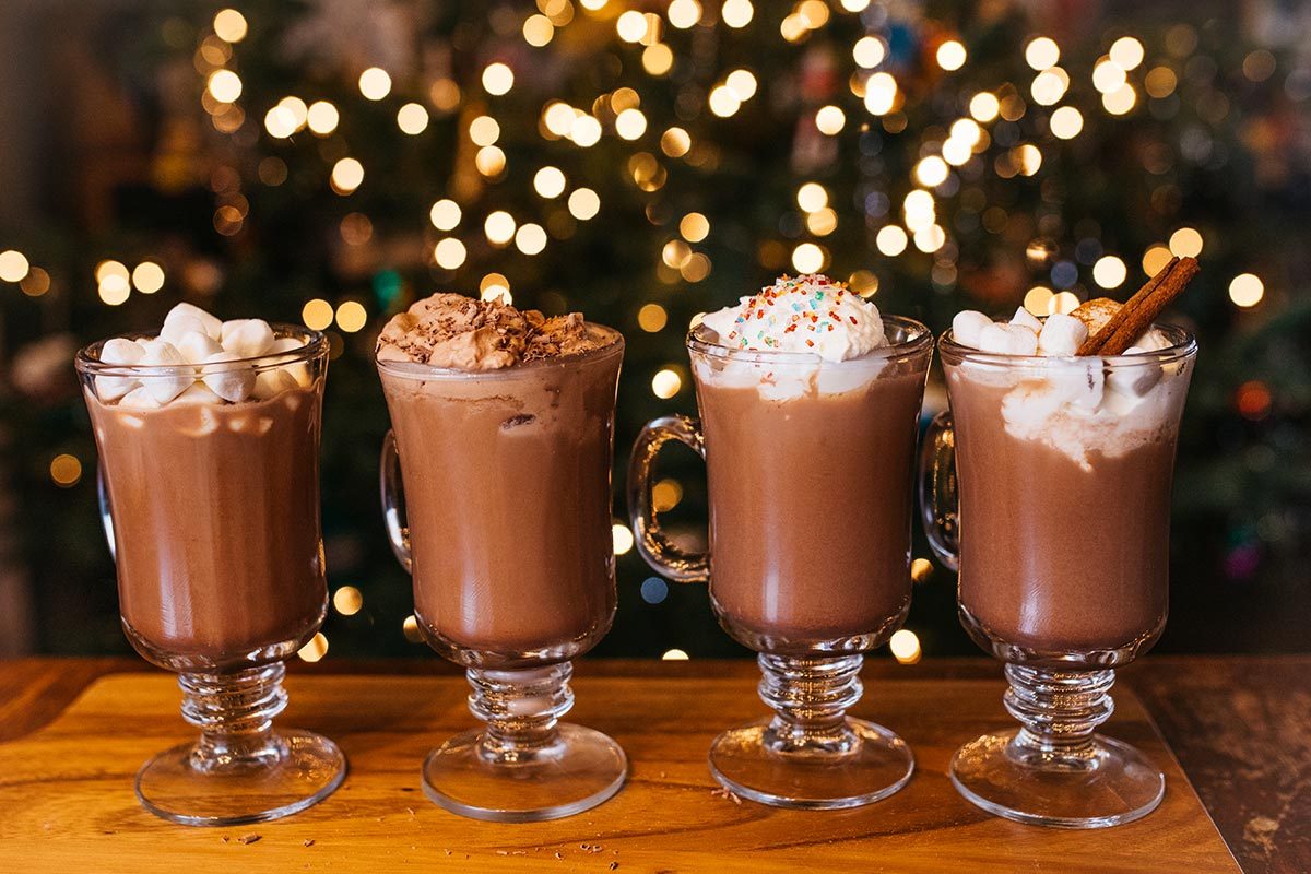 Four glass mugs of hot chocolate with different toppings: marshmallows, chocolate whipped cream, whipped cream and sprinkles, and marshmallows with a cinnamon stick, all on a wooden table with holiday lights in the background