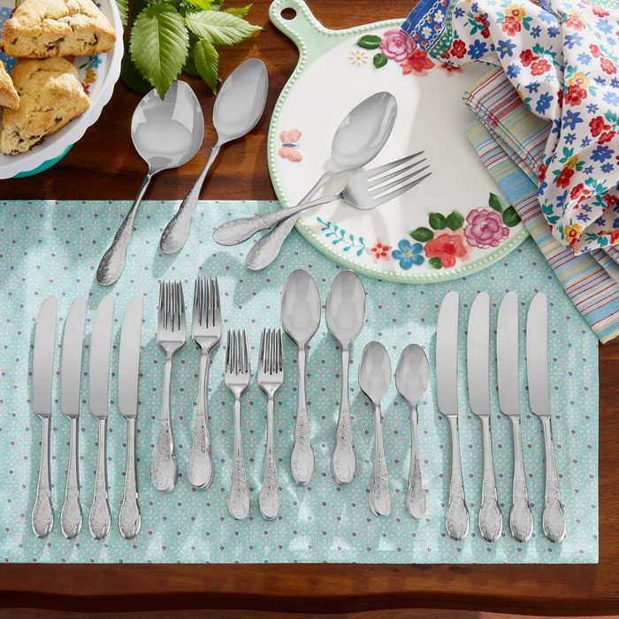 Many pieces of silverware sit on a teal place mat, along with a floral plate and towel, and a bowl of baked goods