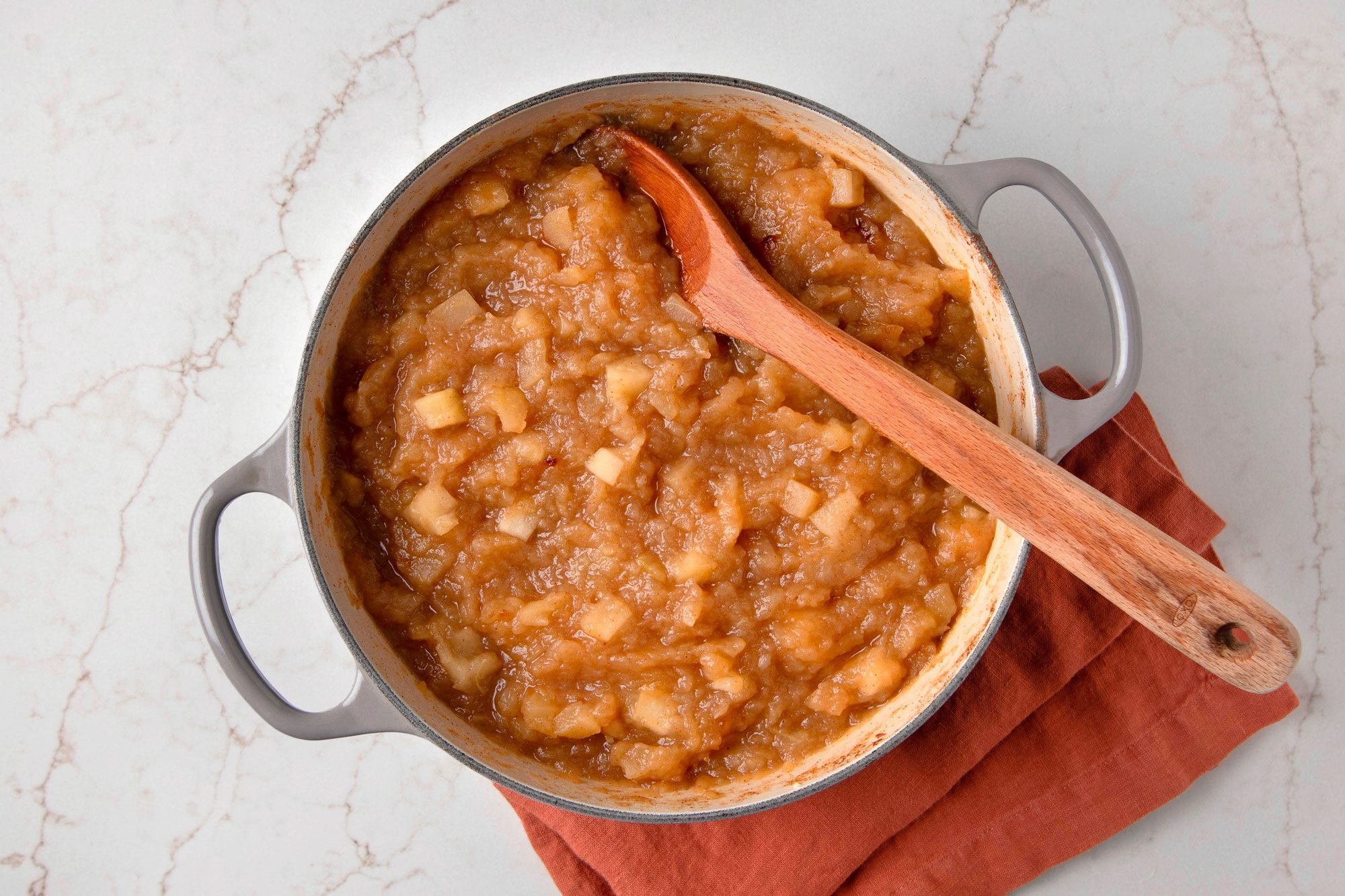 Overhead shot of a pot of applesauce, a wooden spoon, and a maroon kitchen towel, all on a white marble counter