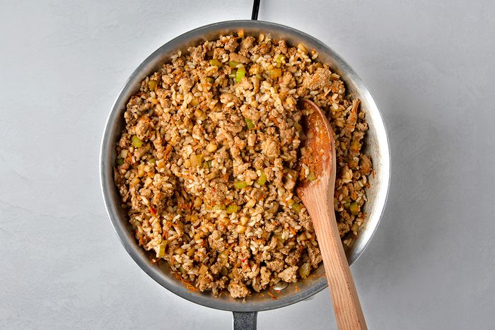 A skillet filled with cooked ground meat, chopped vegetables, and rice, with a wooden spoon resting on the side. The mixture appears seasoned, with visible bits of green bell pepper and a hint of spice.