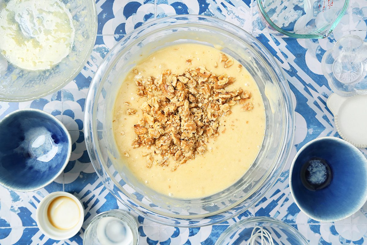 A clear bowl filled with batter and chopped nuts on top sits on a blue and white patterned surface. Surrounding it are various bowls and measuring cups, some with ingredients like flour and cream. Mixing tools are also visible.