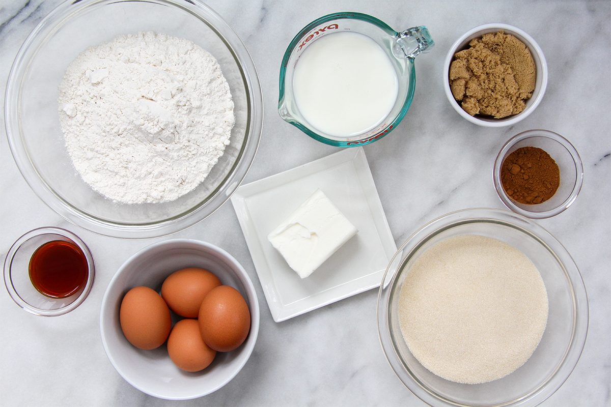 Baking ingredients arranged on a marble surface: bowls of flour, brown sugar, white sugar, and eggs; a square of butter on a plate; a clear measuring cup of milk; small bowls of vanilla extract and cinnamon.