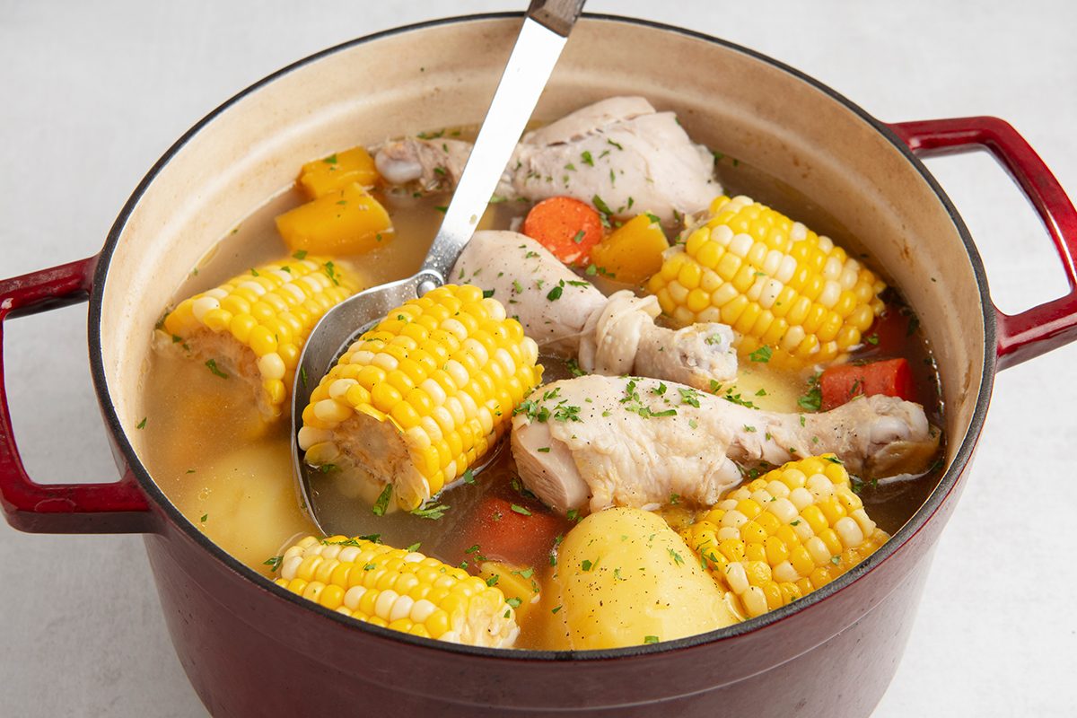 A red pot filled with chicken drumsticks, yellow corn cobs, carrots, and potatoes in a broth. A ladle rests inside, showing chopped parsley garnish on the surface.