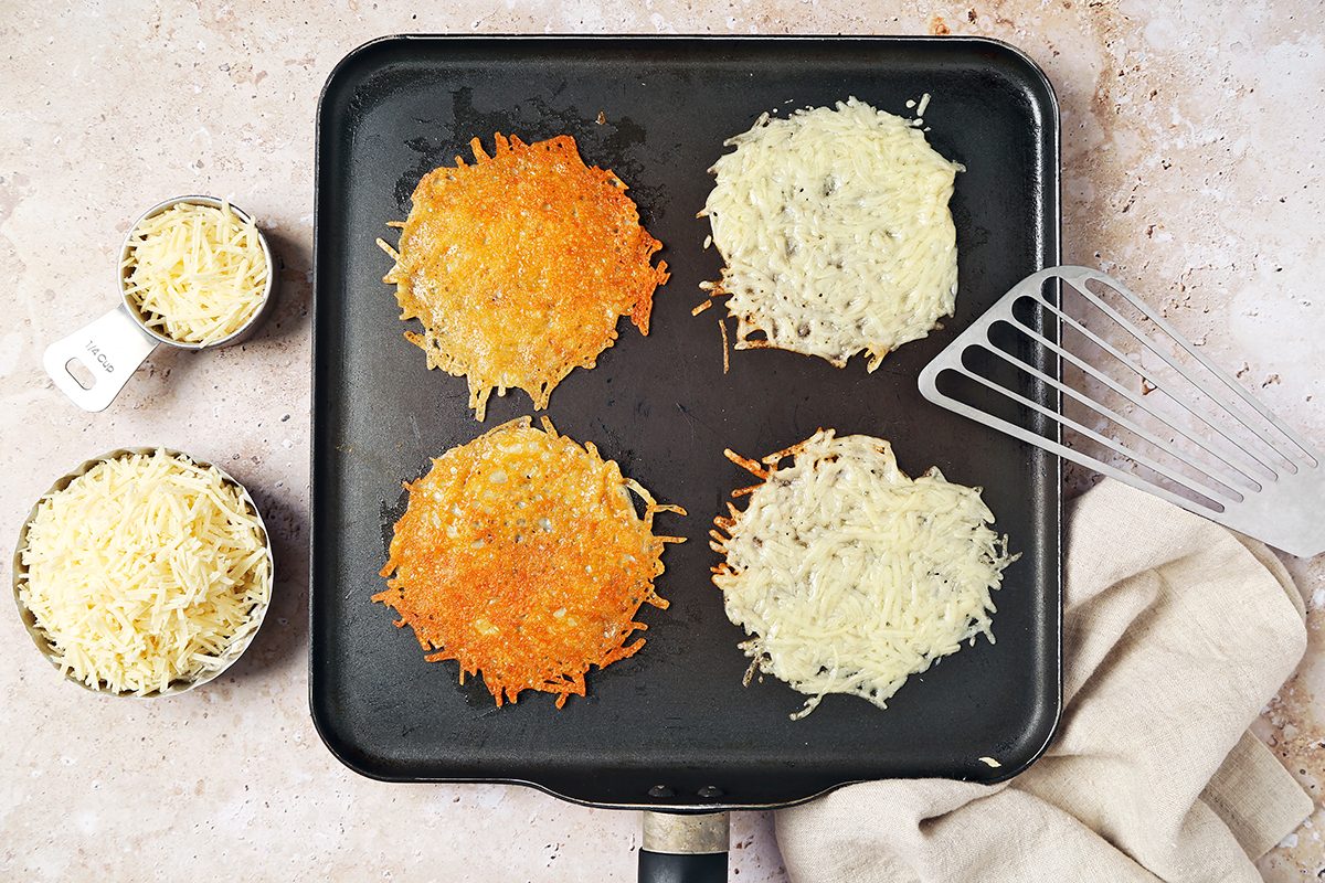 A skillet on a speckled countertop holds four hash browns, two golden brown and two uncooked. Nearby are bowls of grated cheese and a metal spatula resting on a beige cloth.