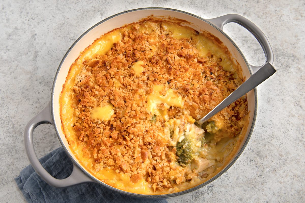 overhead shot of a casserole dish filled with a creamy, cheesy casserole topped with a layer of crispy bread crumbs; the casserole is likely chicken and broccoli, as small pieces of broccoli are visible beneath the bread crumbs; a silver serving spoon lies on top of the casserole dish; the dish is sitting on a gray tablecloth and a gray background