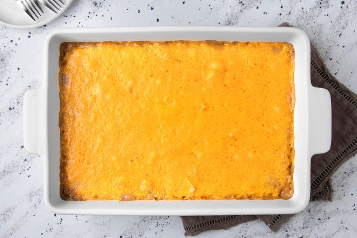 overhead shot of a white baking dish filled with a golden-yellow cheesy casserole; The baking dish is placed on a white surface with a light brown cloth napkin beside it, a white plate with silver forks lying in the top left corner of the image