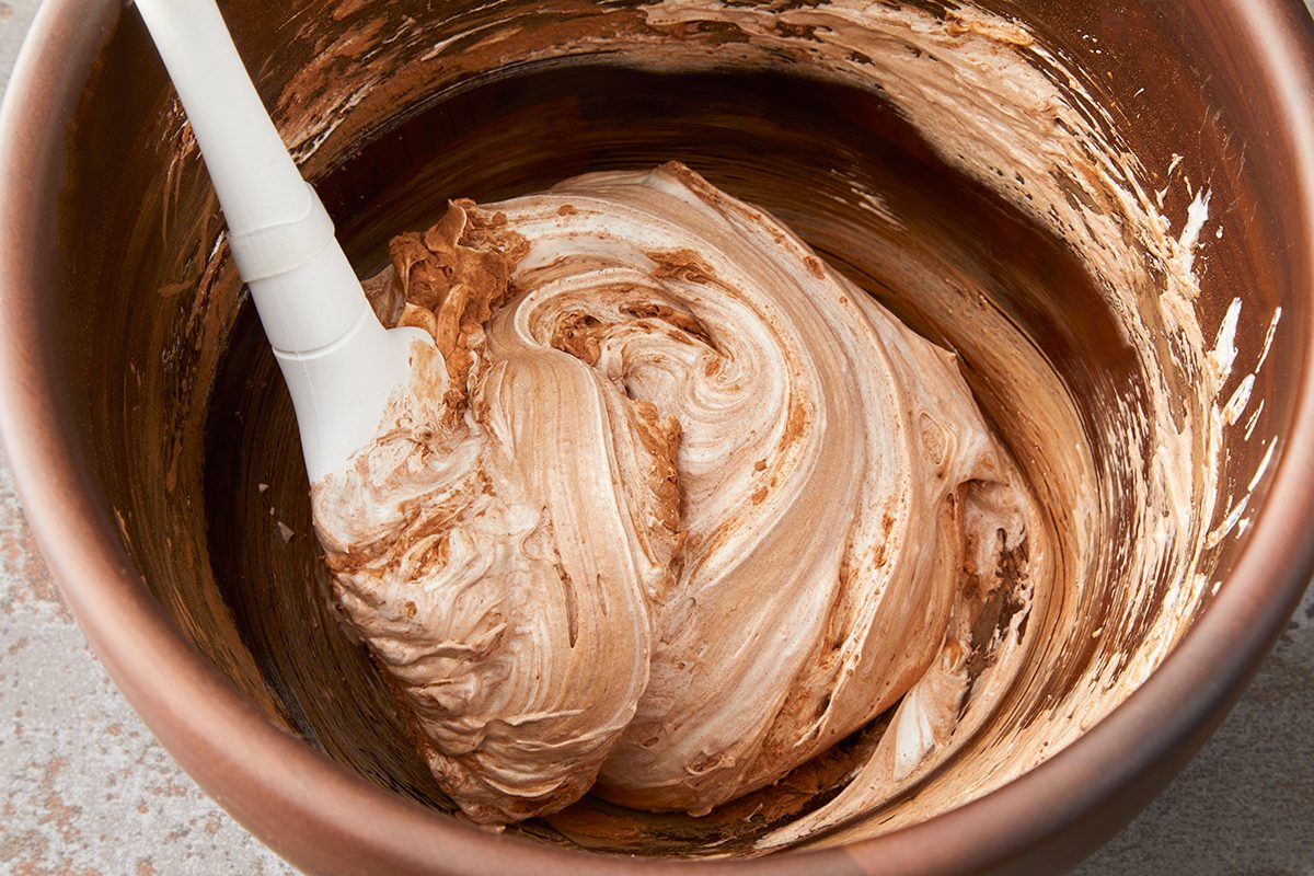 A mixing bowl with swirled chocolate and cream mixture, partially blended with a white spatula resting inside. The mixture has a smooth and creamy texture, displaying light and dark brown tones.