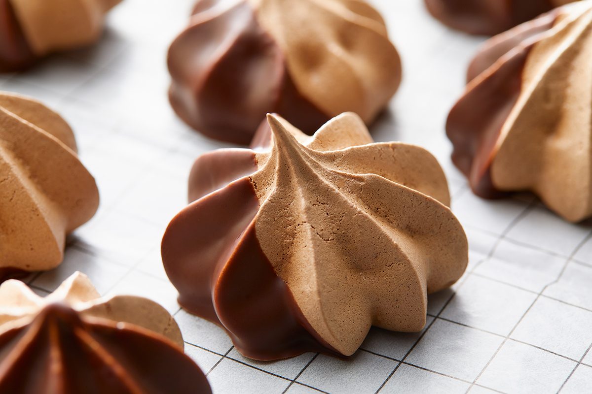 Brown meringue cookies with a glossy chocolate-dipped base are arranged on a white grid-patterned surface. The cookies have a swirled, pointed shape, showcasing a contrast between the matte meringue and shiny chocolate.