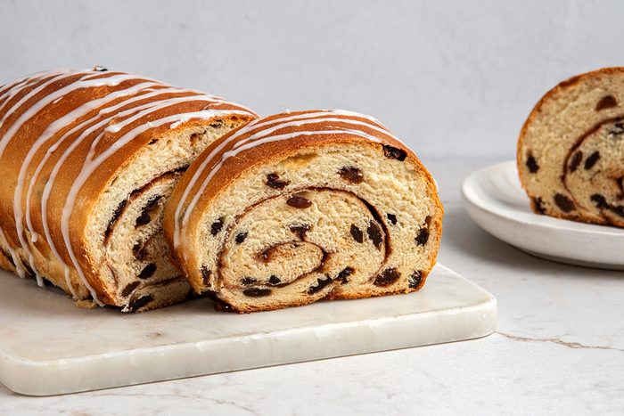 A freshly baked cinnamon raisin bread loaf with a white icing drizzle sits on a marble slab. One slice is cut and placed beside it. Another slice is on a white plate in the background, all set on a light marble surface.