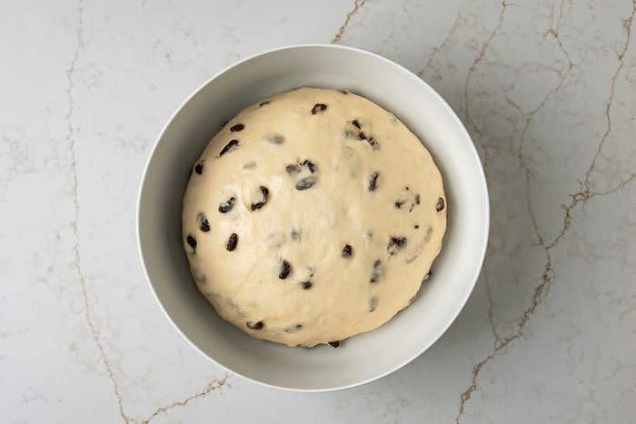A ball of dough with raisins is set in a white mixing bowl, placed on a marbled countertop.