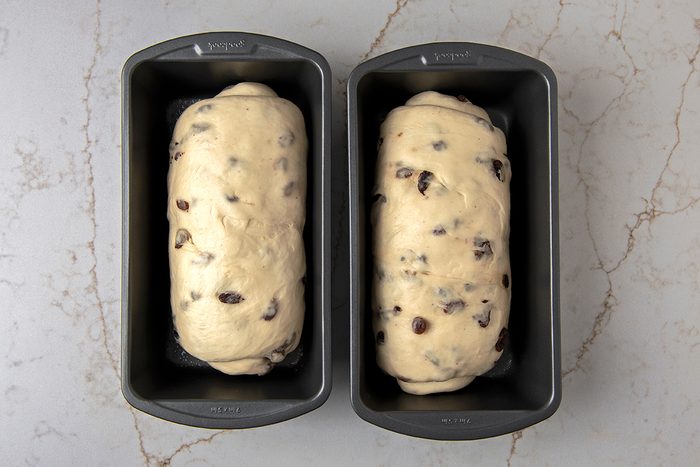 Two loaf pans sit side by side on a marble surface, each containing uncooked dough mixed with raisins. The dough is risen and ready to be baked, with a light, smooth texture visible on top.