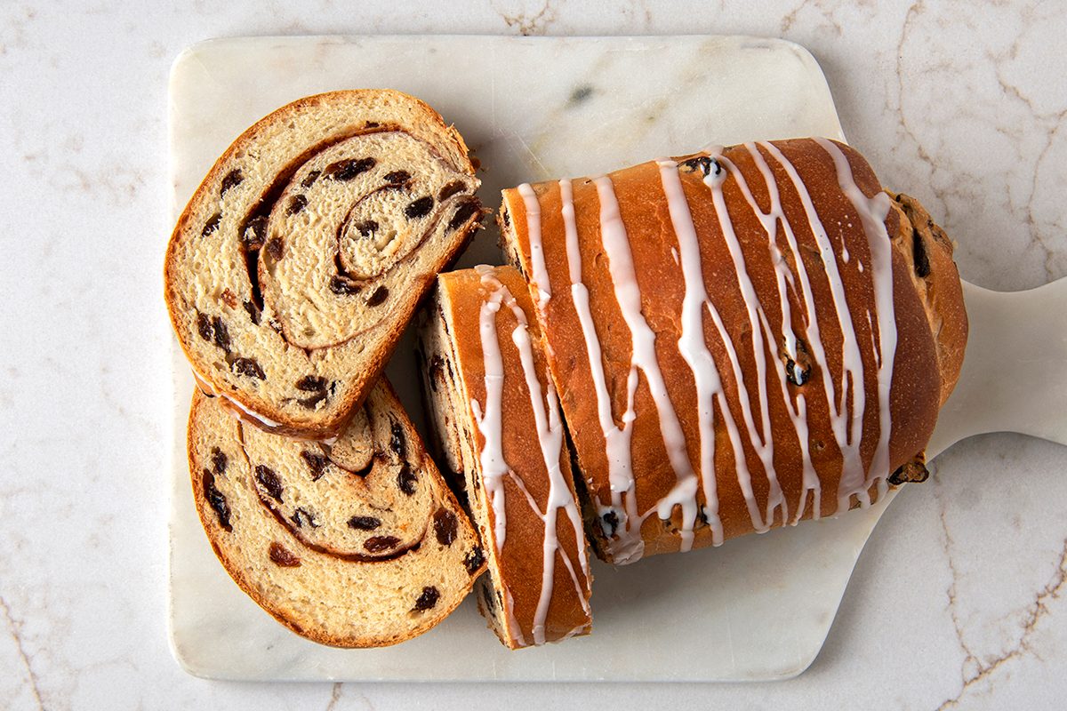 Sliced loaf of cinnamon raisin bread on a marble surface, topped with a drizzle of white icing. The bread is swirled with cinnamon and studded with raisins.