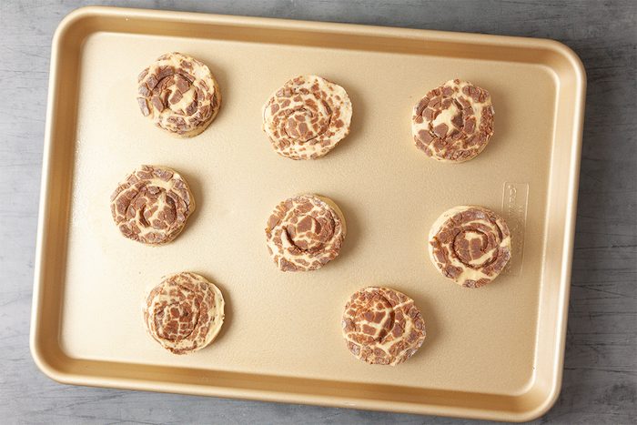 A baking tray with eight uncooked cinnamon rolls evenly spaced on its surface, ready for the oven. The rolls display a swirled pattern with visible cinnamon filling. The tray is placed on a light gray surface.