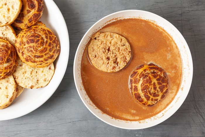 A bowl of rich, brown soup with two pieces of bread soaking in it. Beside the bowl, a plate holds several round, toasted rolls with a patterned crust, likely tiger bread. The background is a gray surface.
