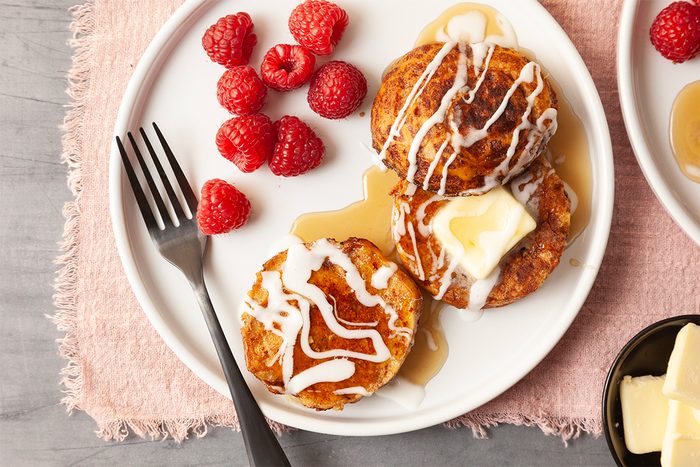 A white plate with two pancakes topped with a pat of butter and white drizzle. Syrup is poured over them. Fresh raspberries are on the side. A fork rests on the plate. A small dish with more butter is nearby, all on a pink cloth.