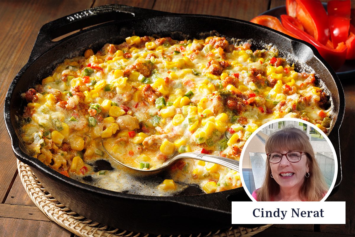 A skillet filled with creamy corn casserole, topped with bits of sausage and green onions. A silver spoon rests inside. A small circular inset shows a person with glasses smiling. Red peppers are visible in the background.