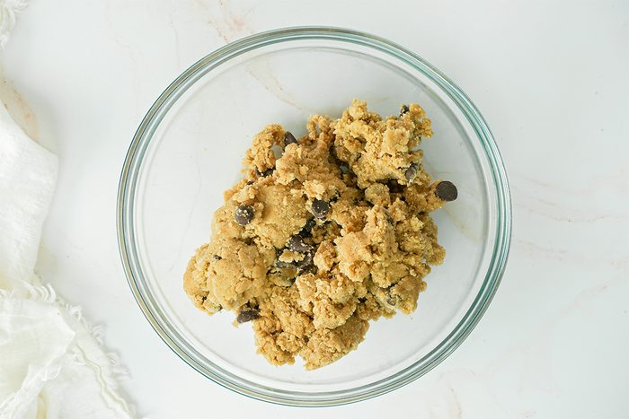 A glass bowl filled with chocolate chip cookie dough on a light-colored surface. A white cloth is partially visible on the left side.