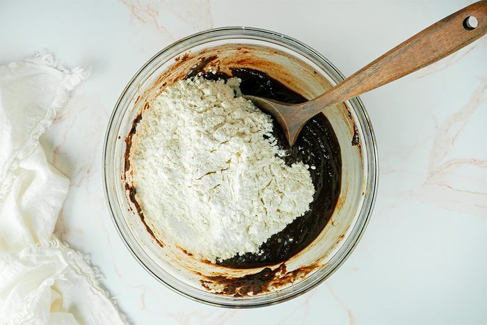 A glass bowl containing a mixture of flour and a dark liquid, likely batter, is being stirred with a wooden spoon. The bowl is placed on a light-colored surface, with a white cloth partially visible to the side.