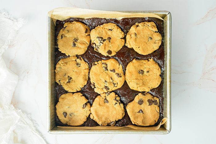 A square baking pan filled with layered dessert bars featuring a dark brownie base topped with nine round cookie dough portions, embedded with chocolate chips. The pan is lined with parchment paper on a light marble surface.