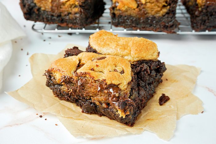 A close-up of a fudgy brownie topped with golden, toasted marshmallows. It's on a piece of parchment paper, with a cooling rack and more brownies in the background. The brownie appears rich and gooey.