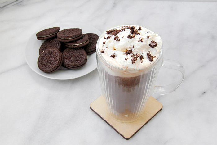 A glass mug of hot chocolate topped with whipped cream and chocolate crumbles sits on a coaster. Next to it is a plate with a stack of chocolate sandwich cookies on a white marble surface.