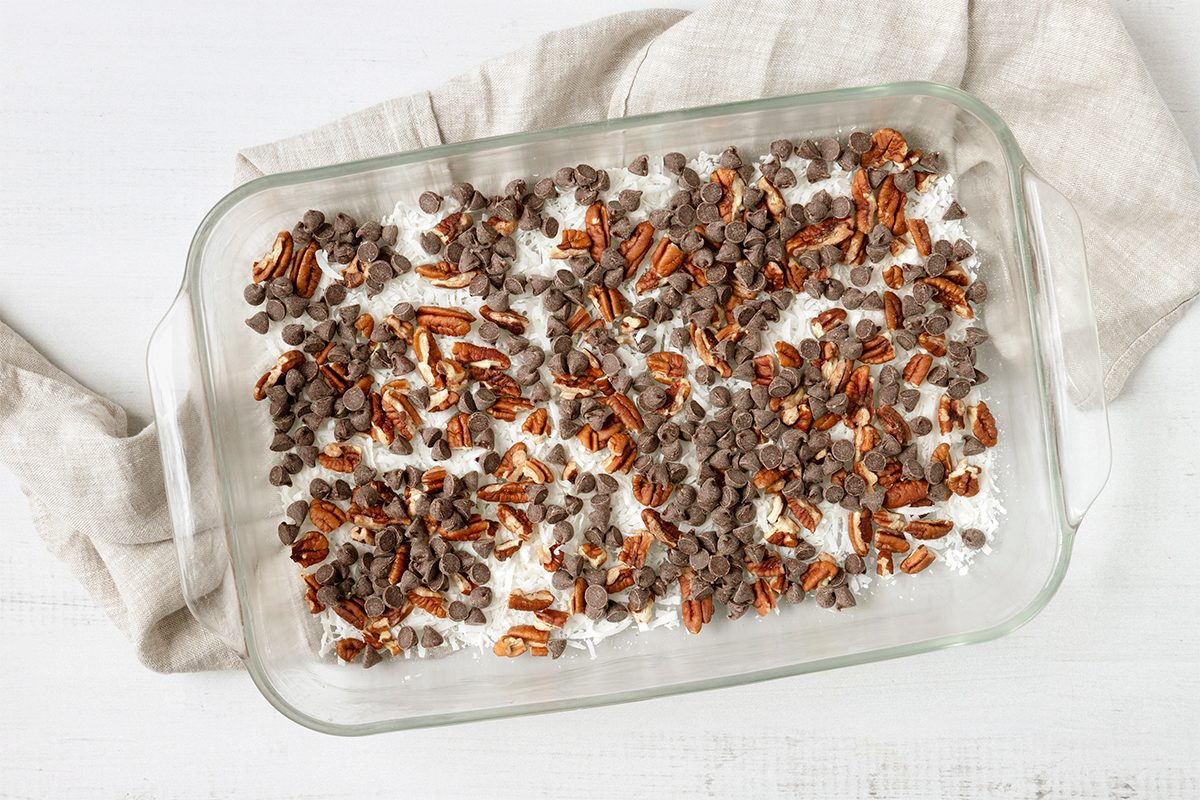 A glass baking dish filled with a dessert topped with chopped pecans and chocolate chips rests on a light-colored surface. A beige cloth is partially tucked under the dish.