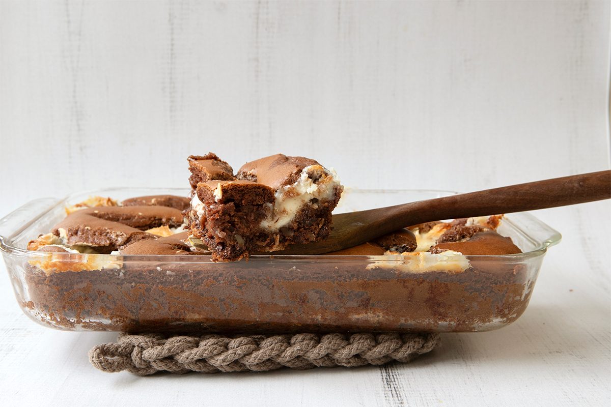 A glass baking dish filled with chocolate and cream dessert sits on a rope trivet. A wooden spoon lifts a portion, showing layers of rich, gooey textures. The background is a simple white surface.