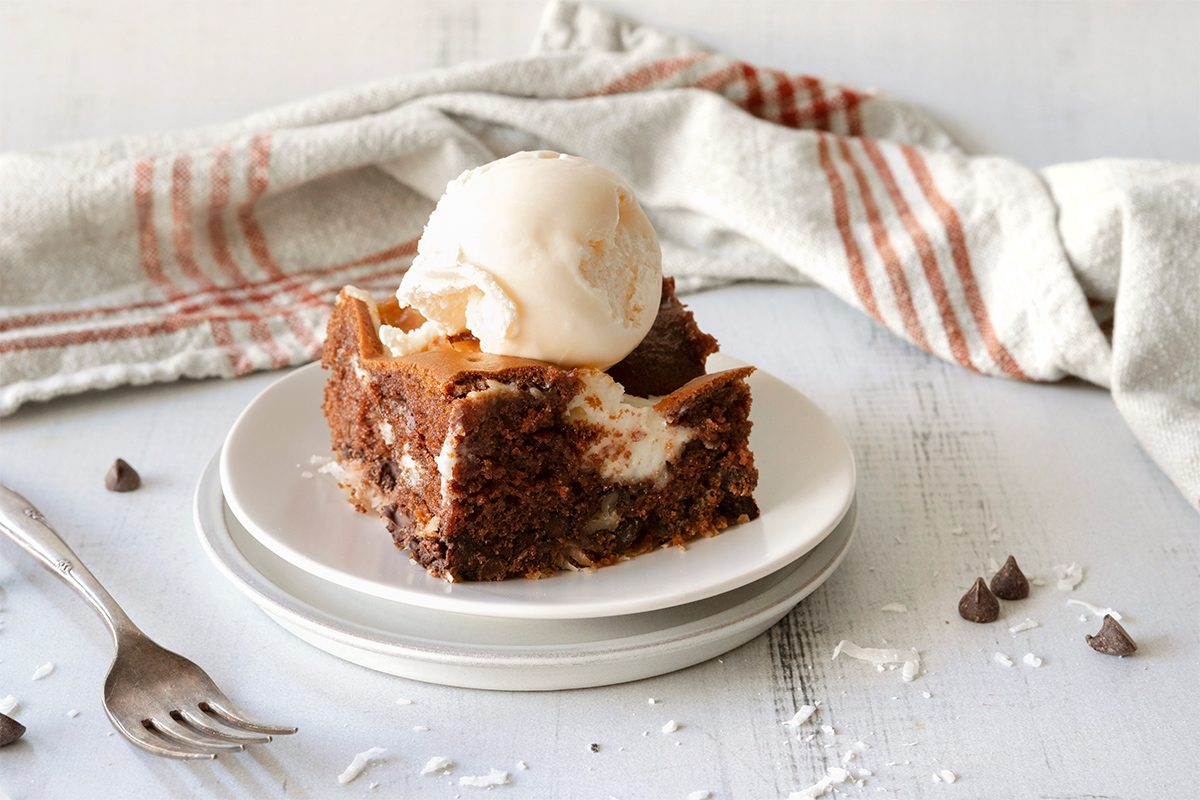 A slice of chocolate brownie with a scoop of vanilla ice cream on top, served on stacked white plates. A fork is placed to the side, and chocolate chips are scattered on a rustic white wooden surface. A striped cloth is in the background.