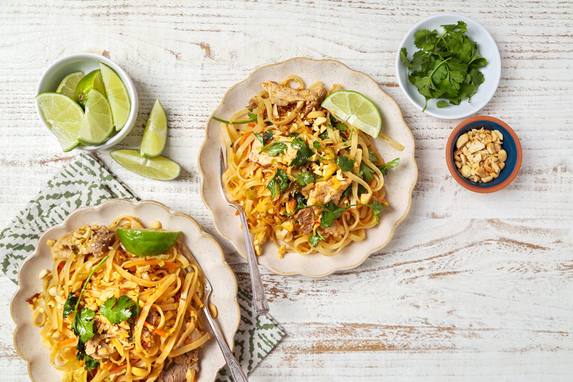 Two plates of Easy Pad Thai, both plates are garnished with fresh cilantro and lime wedges and in a a small bowl of chopped peanuts sits nearby, along with another bowl of fresh cilantro