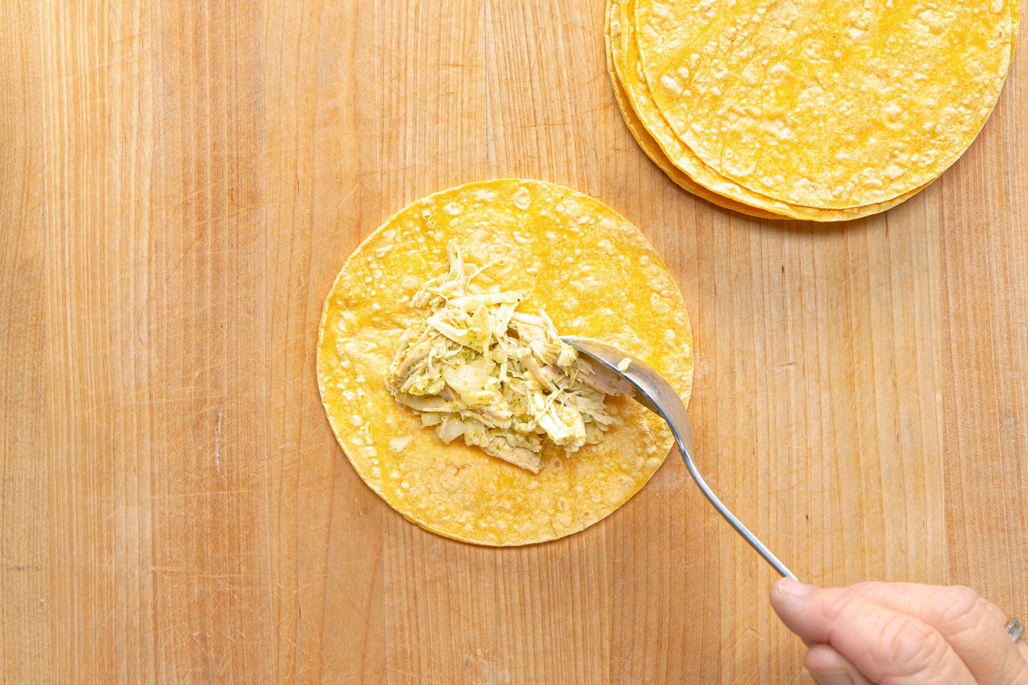 overhead shot of a person preparing a dish on a wooden cutting board; there is a large tortilla placed on the board and a hand is using a spoon to place a generous portion of shredded chicken and green sauce on the tortilla, another pile of smaller tortillas can be seen on the upper right corner of the board