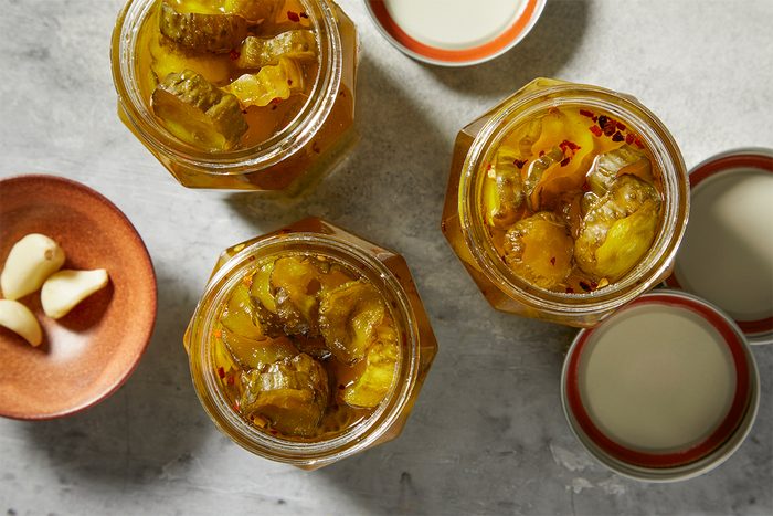 Three jars filled with pickled cucumbers are arranged on a gray surface. Nearby, a small dish holds garlic cloves, and two lids rest beside the jars.