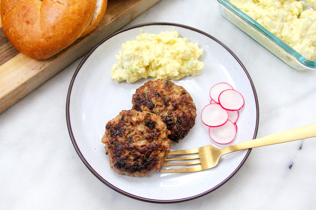 A plate with two browned sausage patties, a scoop of scrambled eggs, and slices of radish. A golden fork is on the plate. A bun is partially visible on a cutting board, with more scrambled eggs in a glass dish nearby.