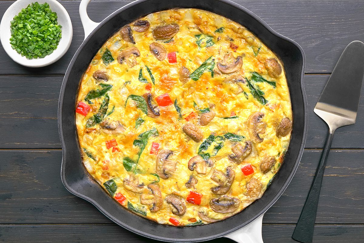A cooked vegetable frittata in a black skillet on a wooden table. The frittata contains visible mushrooms, spinach, and red bell peppers. A small bowl of chopped chives and a spatula are placed beside the skillet.