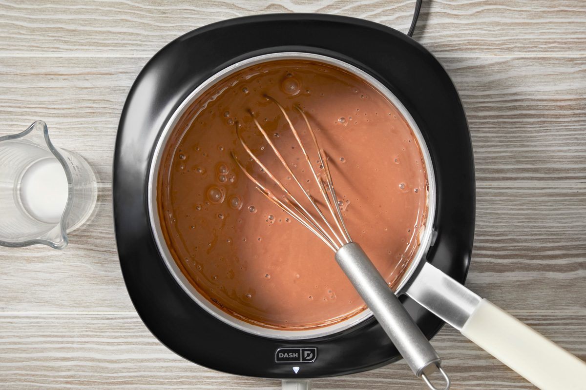 overhead shot of a black electric skillet sits on a white wooden table, a silver whisk lies across the right edge of the image, and a clear measuring cup with liquid sits in the left side of the picture