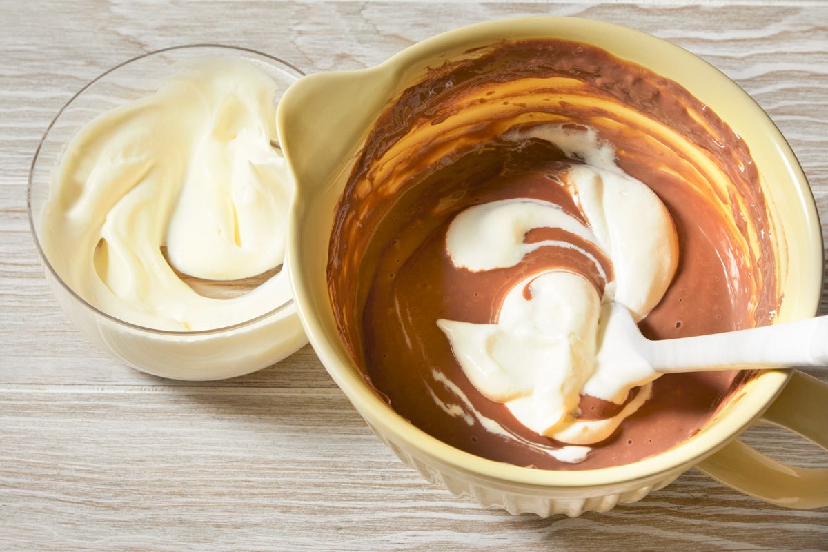 3/4th shot of a bowl of chocolate pudding with whipped cream on top; a clear glass bowl with whipped cream sits next to the pudding bowl; a white spatula is resting on the top of the whipped cream in the pudding bowl; the bowls are both sitting on a light brown wooden surface